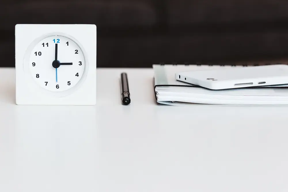 A small square clock, a pen and a closed notebook on a white desk