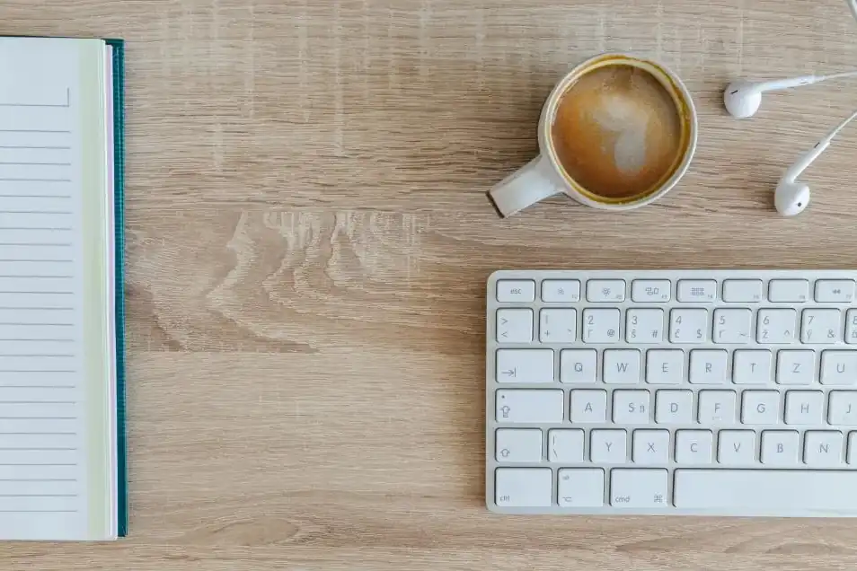 A wooden desk seen from above: notebook, coffee, keyboard and earphones